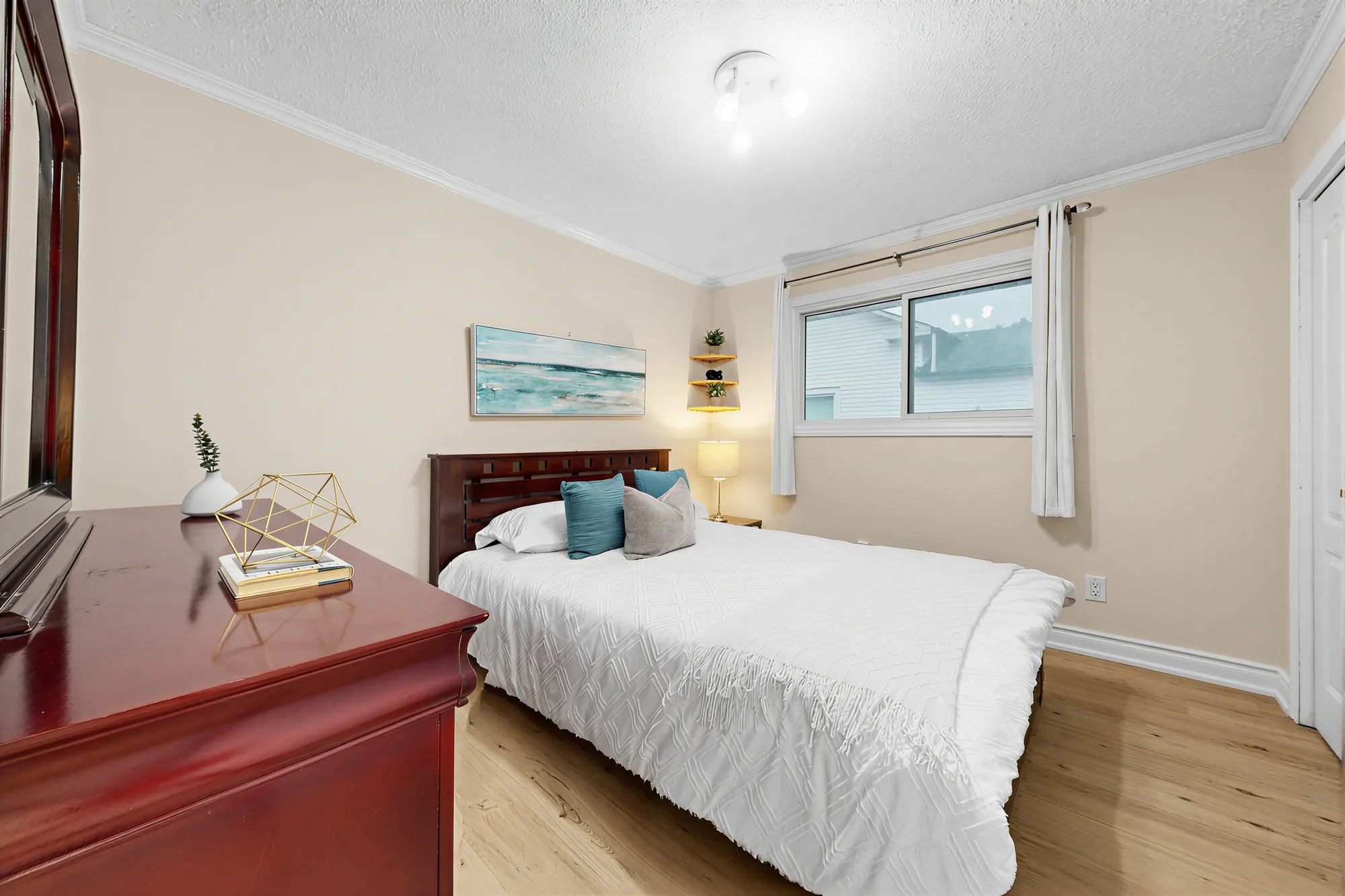 A bedroom with a cherry dresser, a queen bed with white linen, a window with curtains, floating shelves
