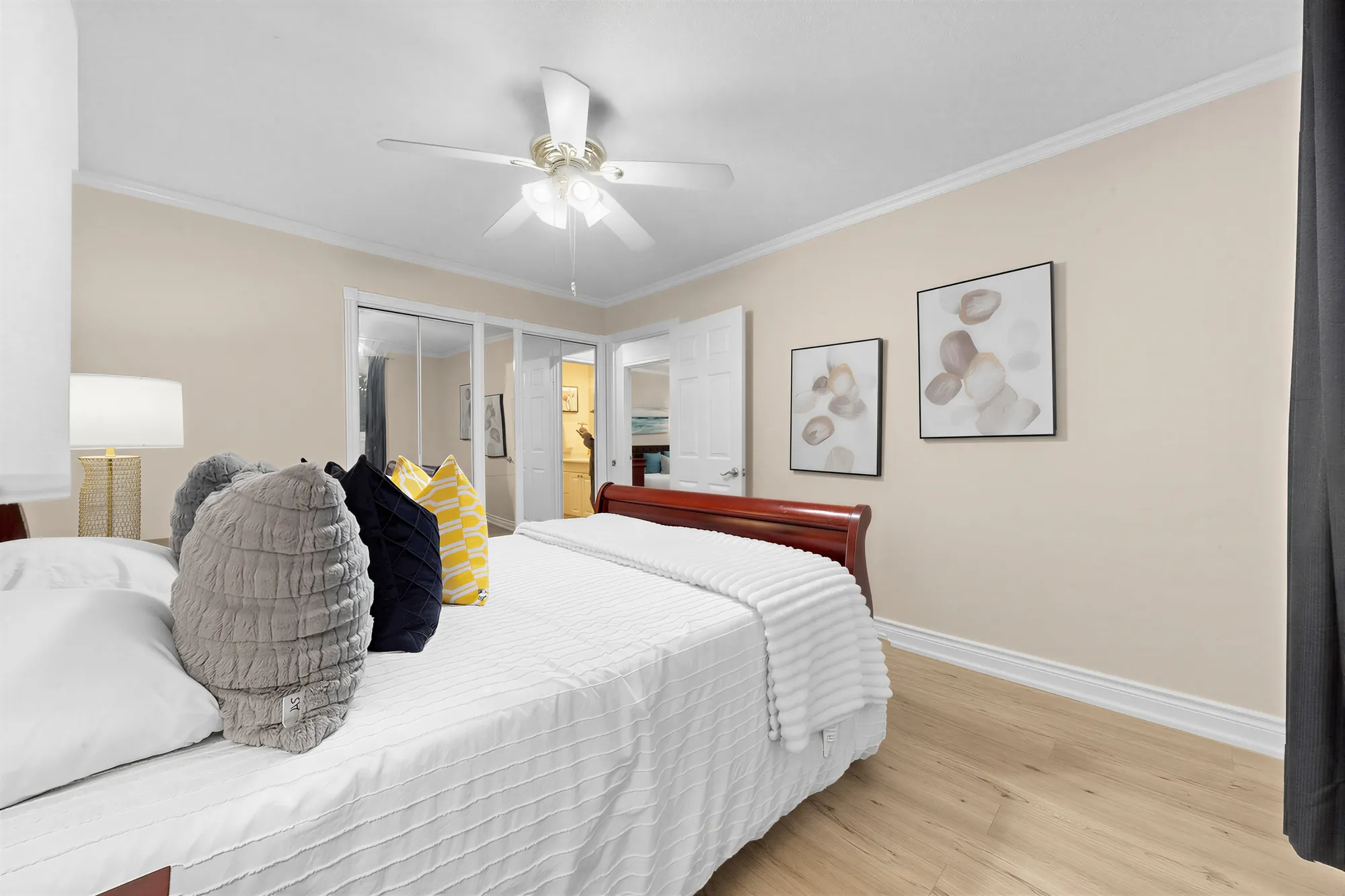 A bedroom with a sleigh bed, a ceiling fan, framed prints and mirrored closet doors, pale laminate floors
