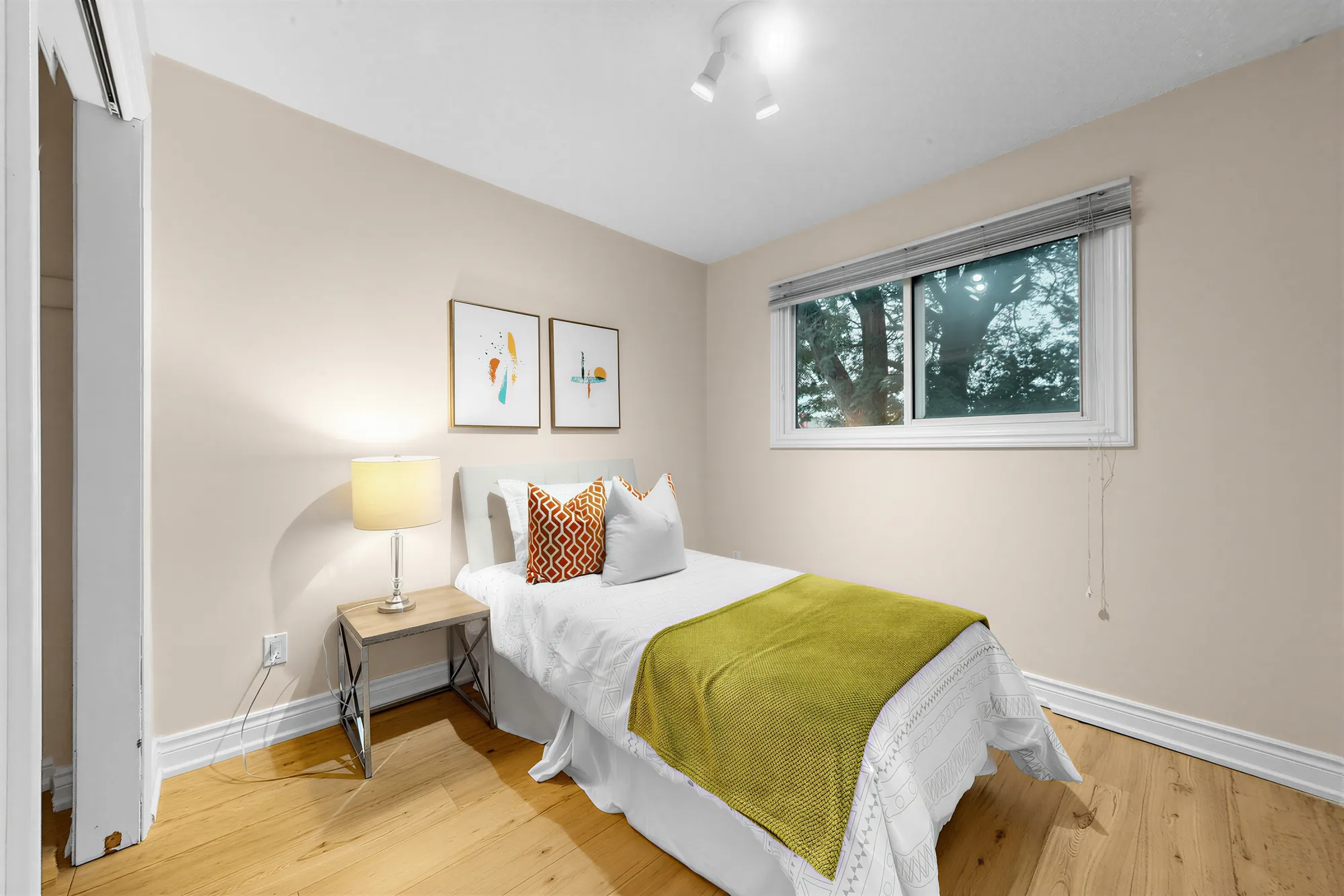 A smaller bedroom: a single bed with a green throw, a night table and lamp, two small prints, a window