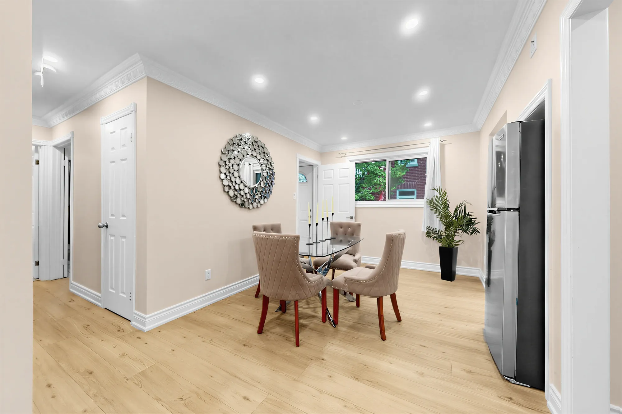 The dining room toward a window and a plant, the fridge at the right, a door at the left