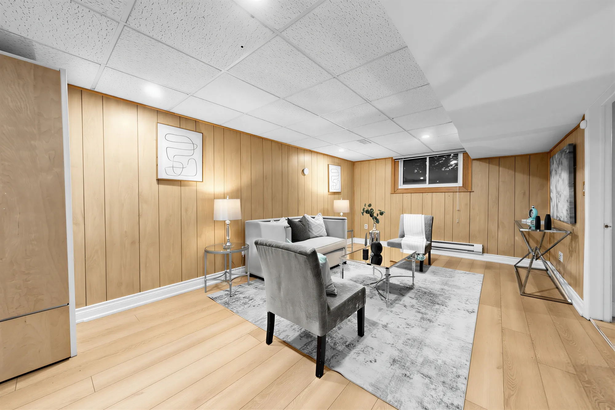 The lower family room: wood-panelled walls, a dropped ceiling with pot lights, a grey sofa and armchairs on a rug, a small window