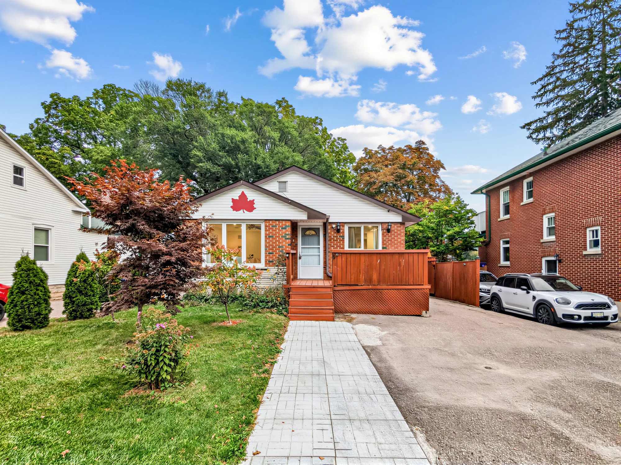 The house from the street: the lawn, the paved walk to the deck, the drive at the right with a car parked, the neighbouring houses on both sides and tall trees behind