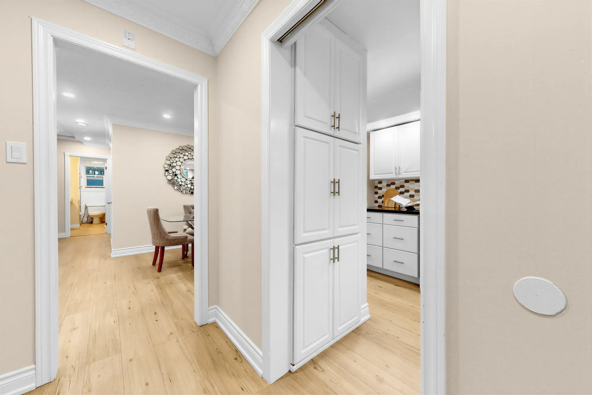 Through a doorway to the dining room and its round mirror, the kitchen&rsquo;s tall white cabinets at the right