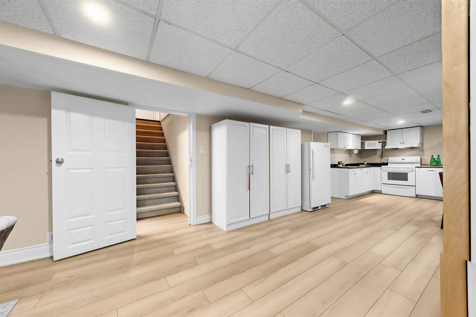 The lower floor toward the stair: a white door at the left, the carpeted stair up, tall white storage cabinets, the kitchen beyond