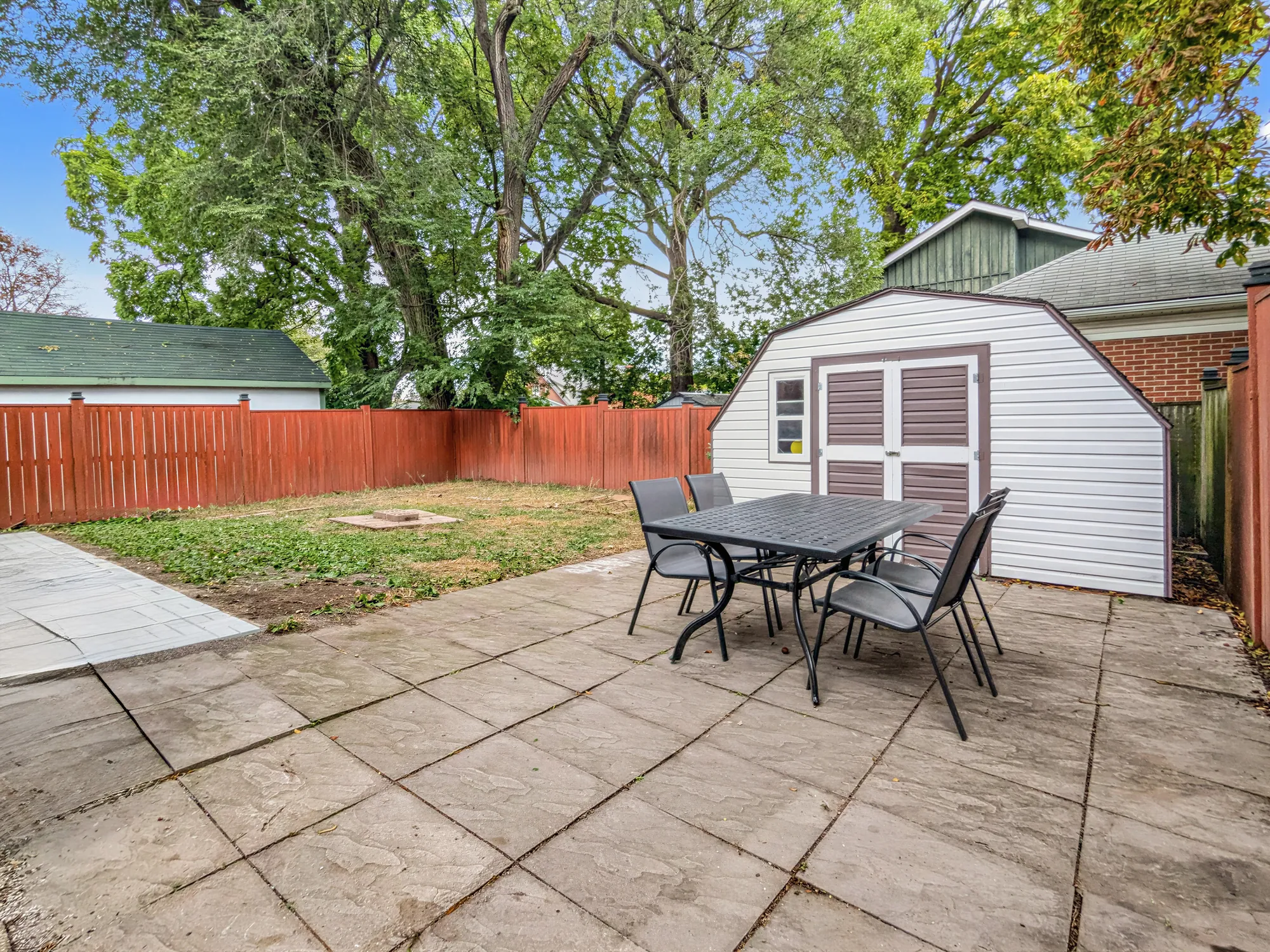 The back: a paved patio with a table and four chairs, a white garden shed, a stained wood fence, the lawn beyond, trees behind