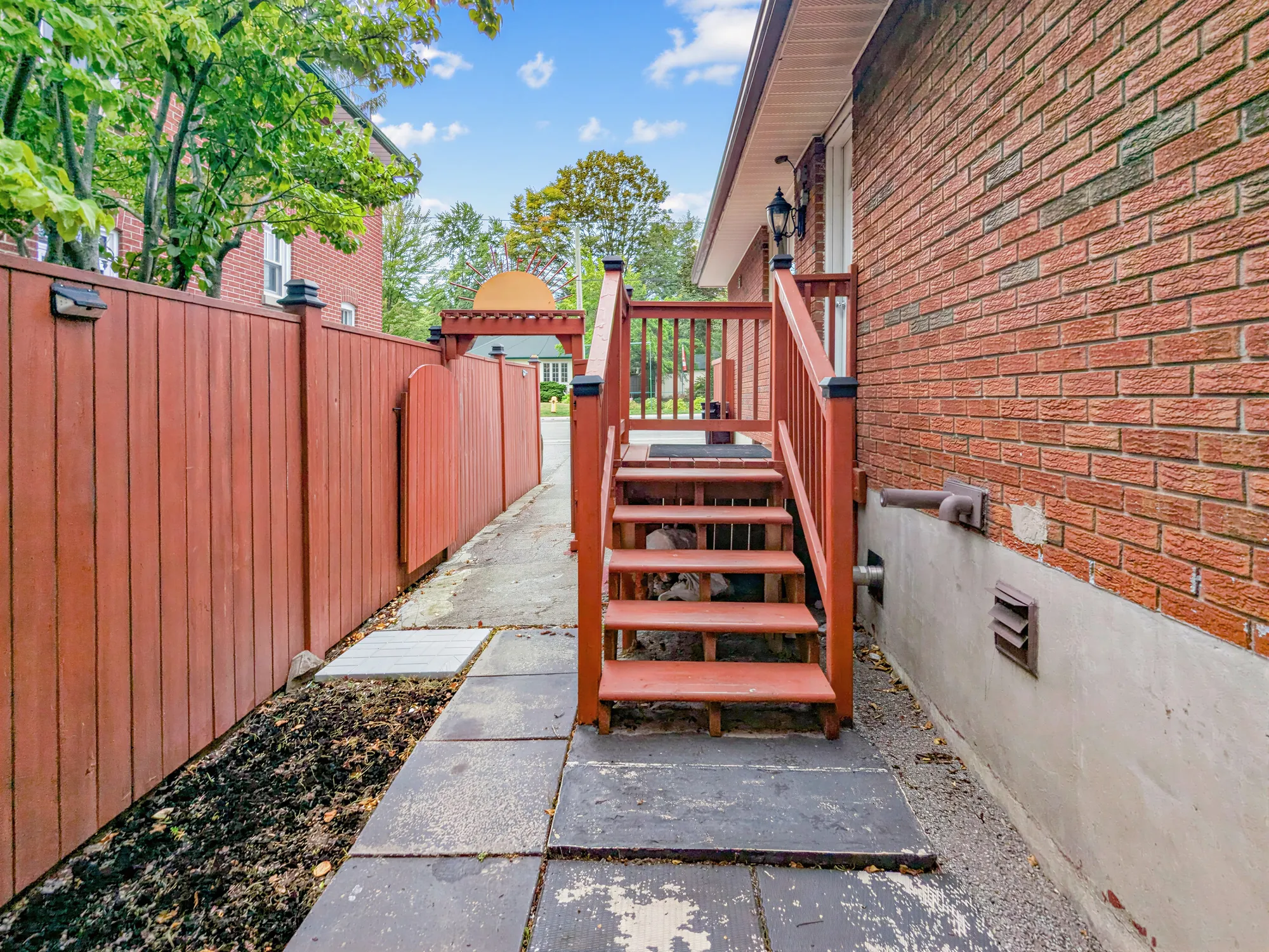 The side path: a paved walk between the brick wall and the stained fence, wooden steps up to the side door, the drive beyond