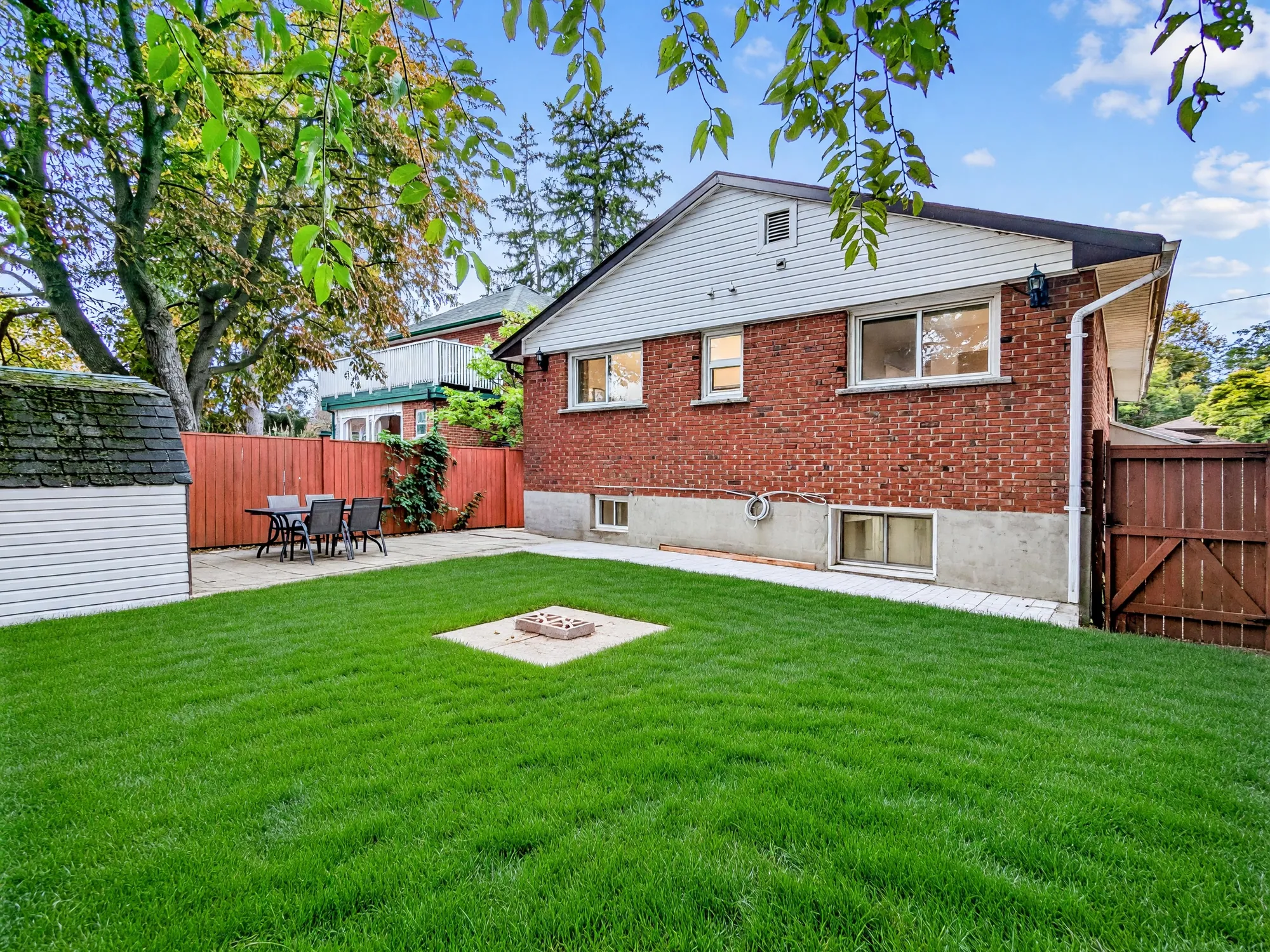The back of the house: red brick under a white-sided gable, two basement windows, a lawn with a small square concrete pad, the shed at the left and a gate at the right